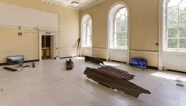 Exhibition image showing a whaler boat in pieces, a scale, video work, and wreck wood value according to the carbon market.